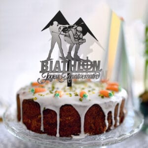 Topper à gâteau biathlon en bois peint blanc, silhouettes de deux athlètes (carabine et ski) avec deux monts en arrière-plan, inscription Joyeux anniversaire en argent miroir